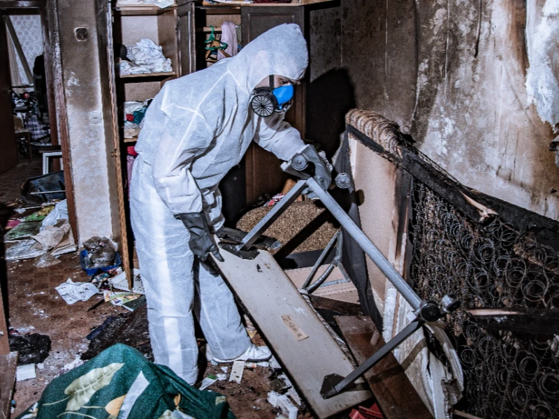 Person in protective suit and mask inspecting fire-damaged furniture in a cluttered room. contents restoration services