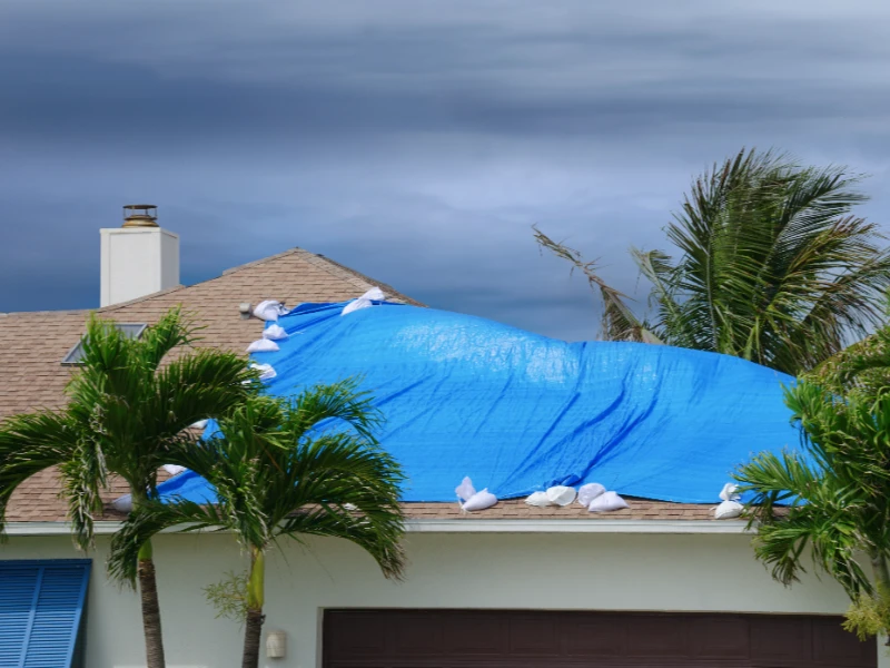 House roof covered with a blue tarp weighted down by sandbags under a cloudy sky. storm damage repair