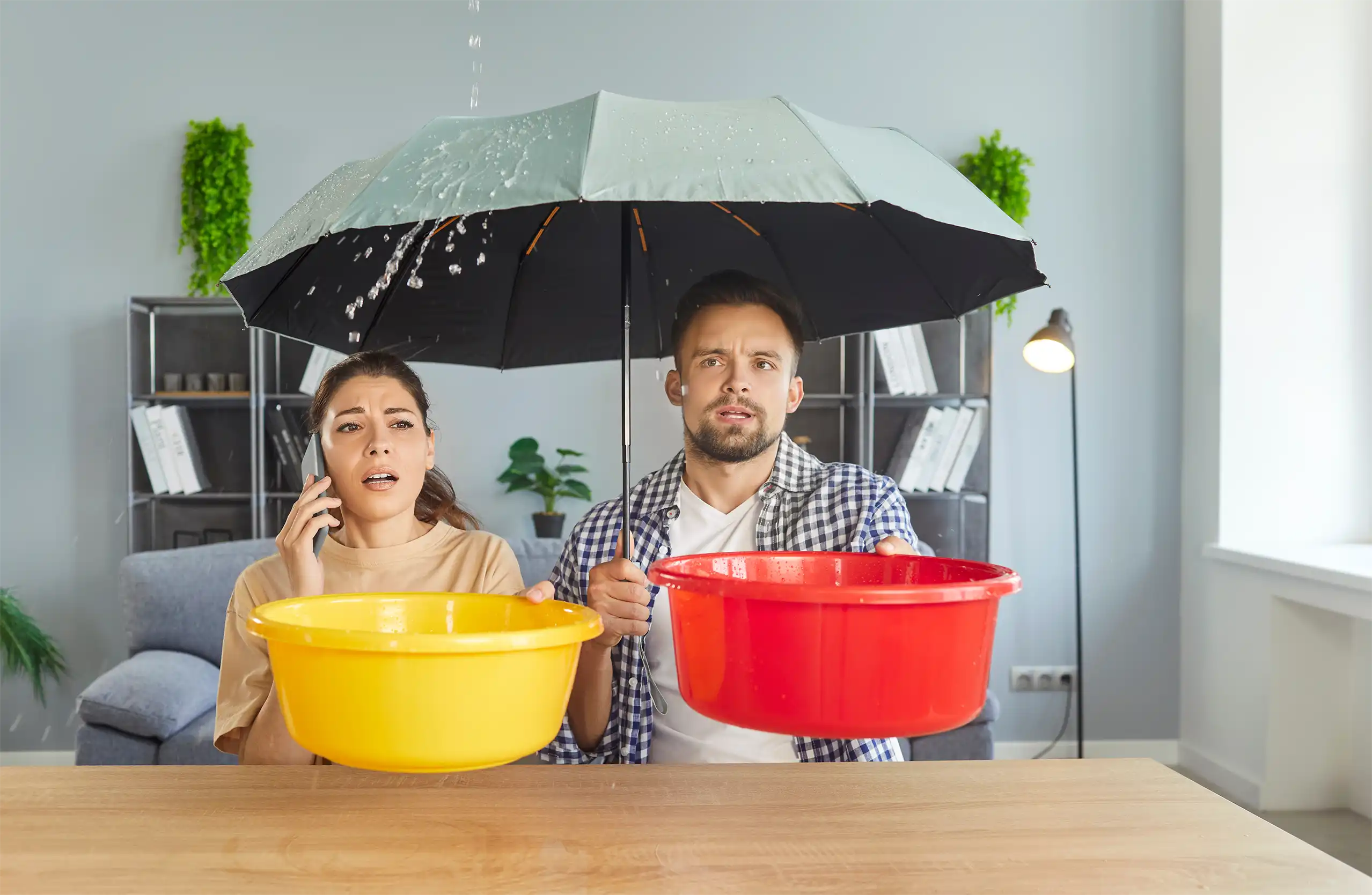 damage restoration services, a man and woman indoors holding an umbrella and bowls to catch water leaking from the ceiling.